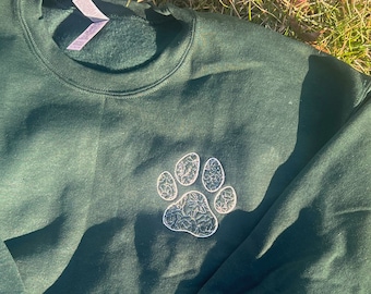 May include: A dark green sweatshirt with a white embroidered paw print design. The paw print is centered on the front of the sweatshirt. The sweatshirt is made of a soft material and has a crew neck.
