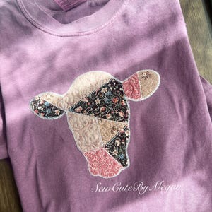 May include: A mauve-colored sweatshirt featuring a patchwork cow head design. The cow head is made of various patterned fabrics, including floral and quilted textures. The text "Sew Cute By Megan" is printed below the cow head.