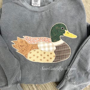 May include: A gray Comfort Colors sweatshirt featuring a patchwork duck design. The duck has a green head, yellow beak, and wings and body made of various patterned fabrics. The text "Sew Cute By Megan" is printed on the bottom right.