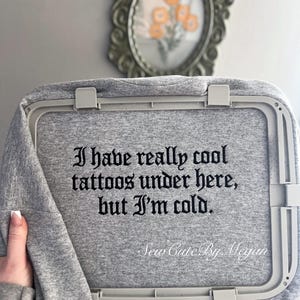 May include: A heather gray sweatshirt secured in a light gray plastic embroidery hoop. The fabric features black embroidered text that reads: I have really cool tattoos under here, but I'm cold. The text is written in a gothic-style font.