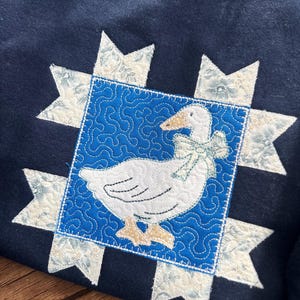 K&ouml;nnte beinhalten: Marineblaues Sweatshirt mit einem gesteppten G&auml;nsemotiv. Die wei&szlig;e Gans hat eine hellblaue Schleife, die in einem blauen Quadrat mit einem Wirbelmuster angeordnet ist. Cremefarbene Sternausbr&uuml;che mit floralem Muster umgeben das Quadrat.
