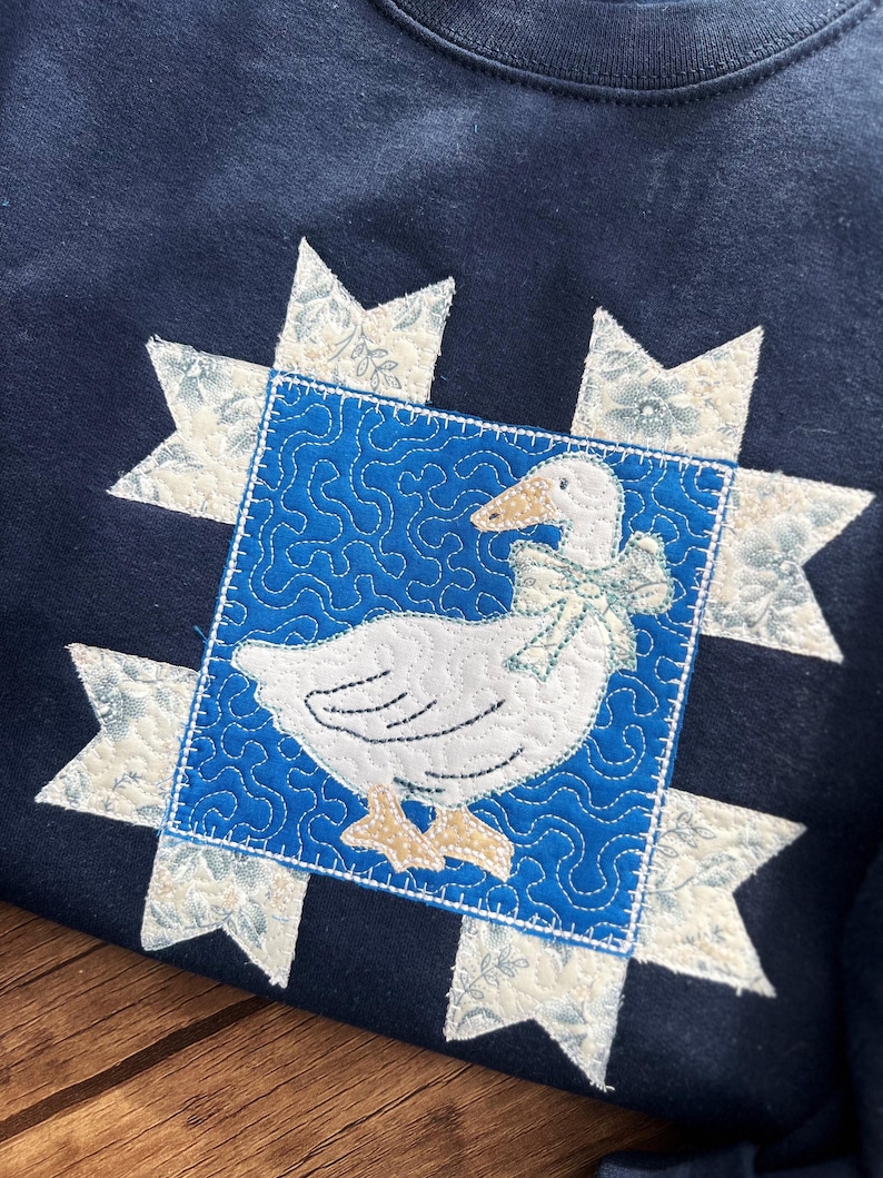 K&ouml;nnte beinhalten: Marineblaues Sweatshirt mit einem gesteppten G&auml;nsemotiv. Die wei&szlig;e Gans hat eine hellblaue Schleife, die in einem blauen Quadrat mit einem Wirbelmuster angeordnet ist. Cremefarbene Sternausbr&uuml;che mit floralem Muster umgeben das Quadrat.