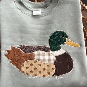 May include: A light green sweatshirt featuring a patchwork duck design. The duck has a paisley green head, a yellow beak, and brown, tan, and plaid patterned body. The text "Sew Cute By Megan" is printed on the bottom.