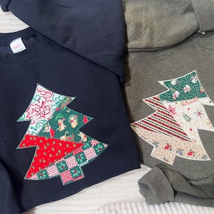 May include: Two sweatshirts, one navy blue and one olive green, each featuring a Christmas tree applique. The trees are constructed from various patterned fabrics in red, green, and cream colors. The sweatshirts are likely made of a soft, comfortable material.