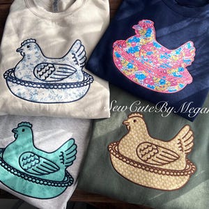 May include: Four sweatshirts with embroidered hen designs. The hen and nest designs feature floral patterns and solid colors, including blue, pink, and teal. The sweatshirts are in cream, navy, gray, and olive green.