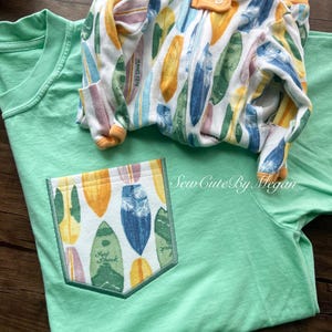 May include: A light green t-shirt with a pocket featuring a surfboard design. A matching onesie with a similar surfboard pattern is folded on top. The onesie has orange trim and buttons. The pocket has the text "Surf Shack".