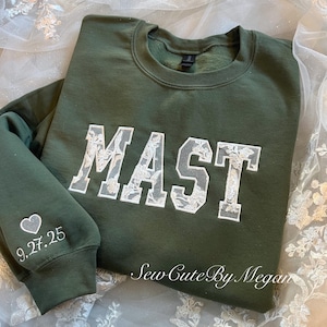 May include: An olive green crewneck sweatshirt featuring the word MAST in large block letters filled with white floral lace applique. The left sleeve displays a small white heart icon above the embroidered date 9.27.25 in white thread.