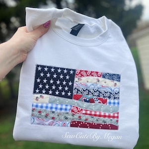 May include: White sweatshirt featuring a patchwork American flag design. The flag includes a navy blue field with white stars and stripes in various patriotic patterns. The text "Sew Cute By Megan" is printed on the bottom right.