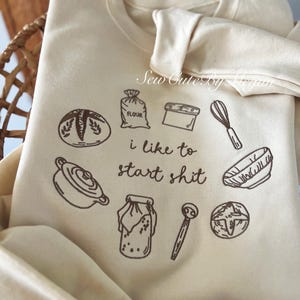 May include: A cream-colored sweatshirt featuring brown embroidered illustrations of baking tools and ingredients, including a bag of flour, a whisk, a bowl, and bread. The text "i like to start shit" is centered among the icons.