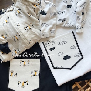 May include: A collection of baby clothing including a cream-colored zip-up sleeper with a tiger face pattern, a white long-sleeve shirt with a black cloud print, and a black shirt with a tiger face pocket. The text Sew Cute By Megan is visible.
