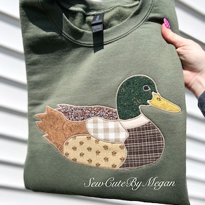 May include: An olive green crewneck sweatshirt featuring a large mallard duck appliqué made from various patterned fabric patches in shades of brown, tan, and green. The text Sew Cute By Megan is embroidered in white script at the bottom right.