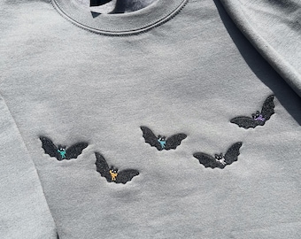May include: Gray sweatshirt with embroidered black bats. Each bat has colorful accents in teal, yellow, and purple. The sweatshirt is made of a soft material and has a crew neck.
