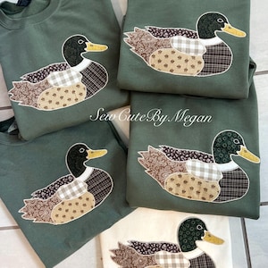 May include: Several olive green sweatshirts and one cream-colored sweatshirt, each adorned with a detailed duck applique. The duck features a black head, yellow beak, and wings and body made of patterned fabrics. The text "Sew Cute By Megan" is visible.