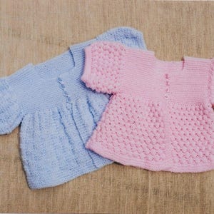May include: Two hand-knitted baby sweaters. One is light blue, the other is pink. Both have a round neckline, short sleeves, and a button closure down the front. The sweaters are made of soft yarn and have a textured stitch pattern.