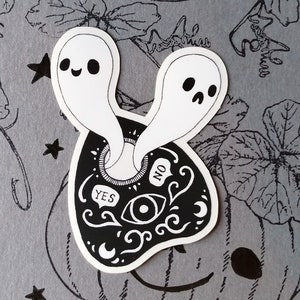 May include: Black and white sticker of a Ouija board with two ghosts peeking out of the opening. The board has the words "YES" and "NO" written on it.