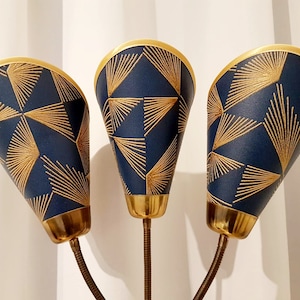 Op de afbeelding: Drie blauwe lampenkappen met gouden rand en een geometrisch patroon van gouden lijnen. De lampenkappen zijn bevestigd aan een gouden metalen basis.