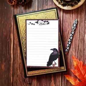 May include: A black and gold journal with a white notepad featuring a black raven illustration and the word "Notes" at the top. The notepad is on a wooden surface with a cup of tea and a fall leaf.