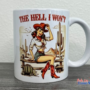 May include: White ceramic mug with a colorful illustration of a woman wearing a cowboy hat and boots, sitting on a fence with cacti in the background. The text "THE HELL I WON'T" is printed on the mug.