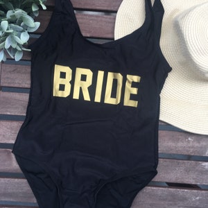 May include: Black one-piece swimsuit with the word "BRIDE" in gold block letters across the front. A beige sun hat and greenery are in the background. The swimsuit has a scoop neck and spaghetti straps.