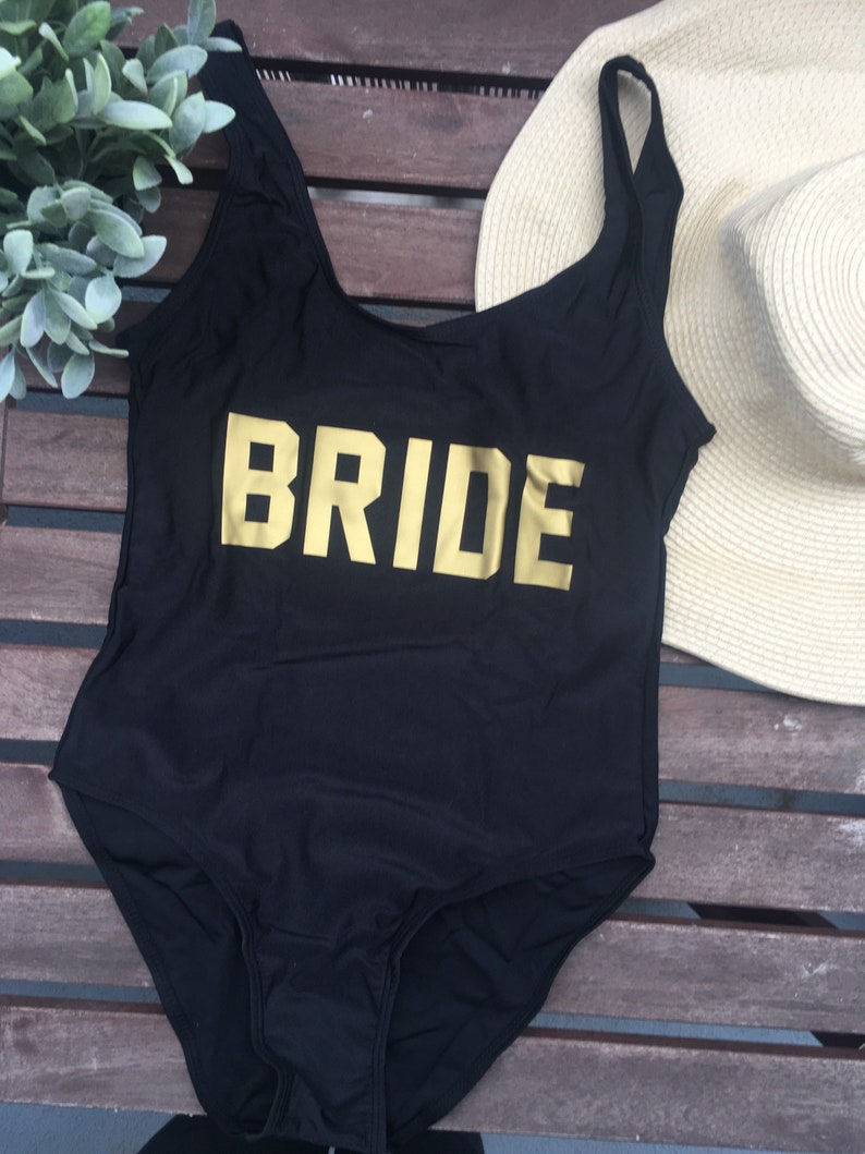 May include: Black one-piece swimsuit with the word "BRIDE" in gold block letters across the front. A beige sun hat and greenery are in the background. The swimsuit has a scoop neck and spaghetti straps.