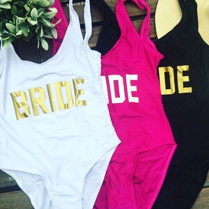 May include: White, pink, and black swimsuits with gold lettering that spells out "BRIDE" and "DE". The swimsuits are displayed on a wooden surface, with a straw hat and greenery in the background. The swimsuits are one-piece.