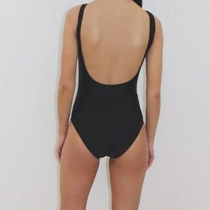 May include: A black one-piece swimsuit with a scoop back and thin straps. The swimsuit is a solid black color and appears to be made of a smooth fabric. The model is standing in front of a white background.