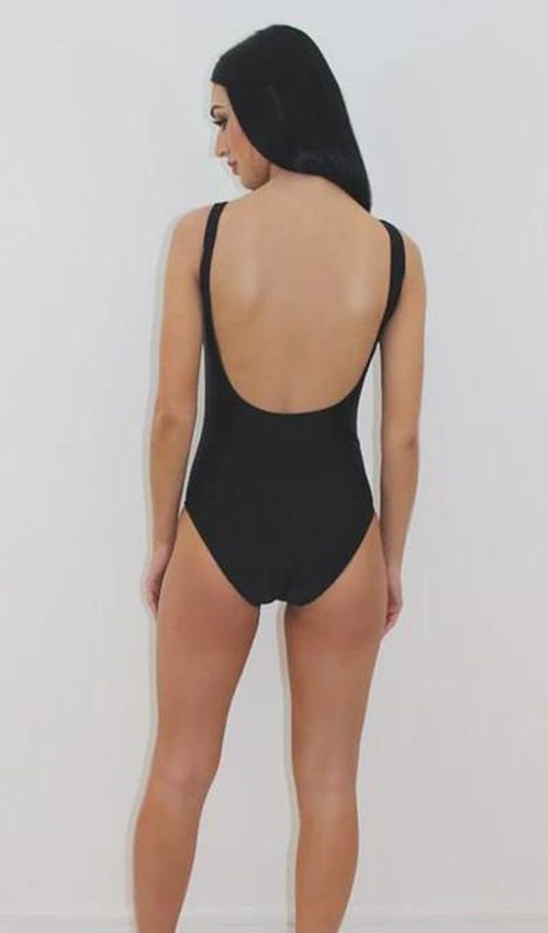 May include: A black one-piece swimsuit with a scoop back and thin straps. The swimsuit is a solid black color and appears to be made of a smooth fabric. The model is standing in front of a white background.