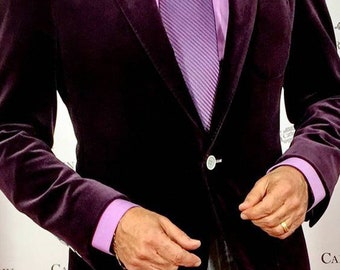 Puede incluir: Un hombre con un blazer de terciopelo morado con una corbata de rayas moradas claras y un pañuelo de bolsillo blanco. También lleva una camisa de vestir morada clara y pantalones de rayas grises.