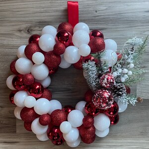 K&ouml;nnte beinhalten: Ein Weihnachtskranz aus roten und wei&szlig;en Kugeln. Der Kranz ist mit einem Tannenzweig und wei&szlig;en Beeren verziert.