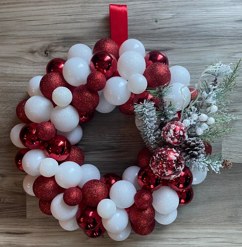 K&ouml;nnte beinhalten: Ein Weihnachtskranz aus roten und wei&szlig;en Kugeln. Der Kranz ist mit einem Tannenzweig und wei&szlig;en Beeren verziert.