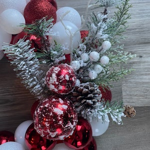 K&ouml;nnte beinhalten: Ein Weihnachtskranz aus roten und wei&szlig;en Kugeln, Tannenzweigen und wei&szlig;en Beeren. Der Kranz ist mit Kunst Schnee verziert.
