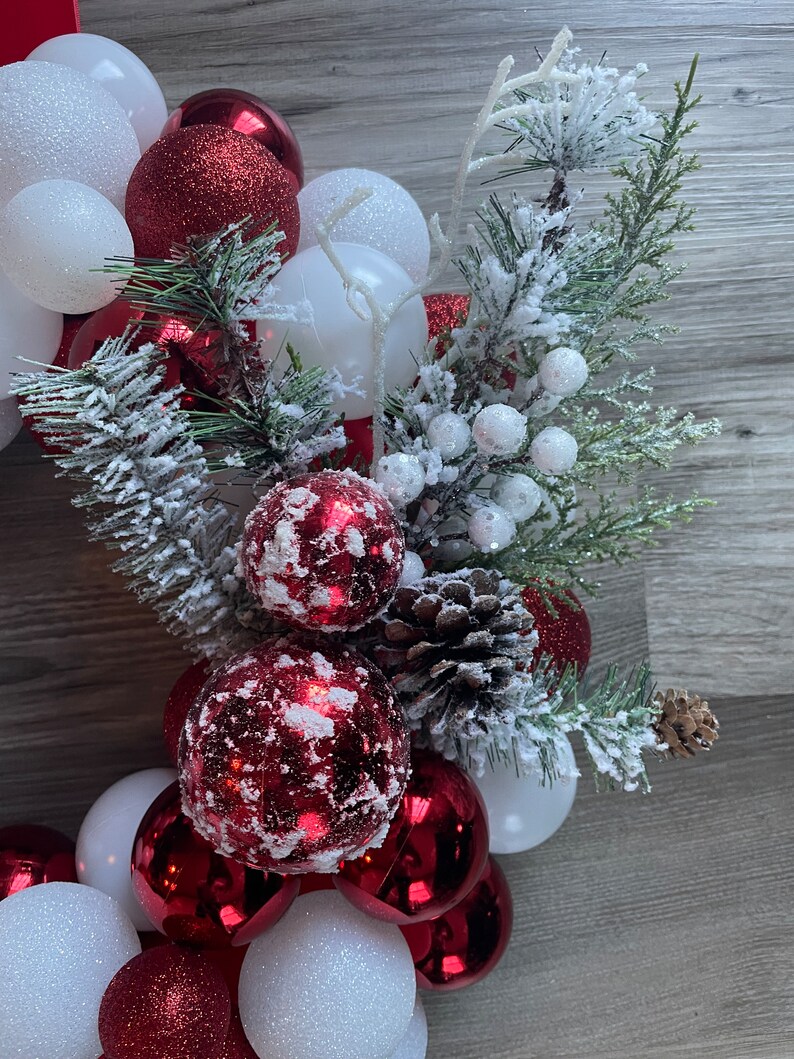 K&ouml;nnte beinhalten: Ein Weihnachtskranz aus roten und wei&szlig;en Kugeln, Tannenzweigen und wei&szlig;en Beeren. Der Kranz ist mit Kunst Schnee verziert.