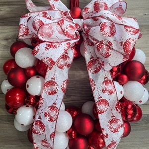 K&ouml;nnte beinhalten: Ein Weihnachtskranz, der mit roten und wei&szlig;en Kugeln und einem wei&szlig;en Band mit rotem Schneeflockenmuster verziert ist. Das Band ist oben am Kranz zu einer gro&szlig;en Schleife gebunden.