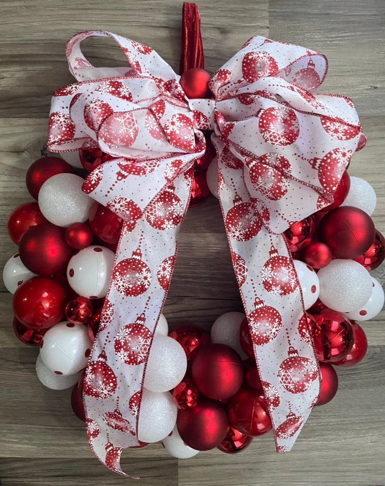 K&ouml;nnte beinhalten: Ein Weihnachtskranz, der mit roten und wei&szlig;en Kugeln und einem wei&szlig;en Band mit rotem Schneeflockenmuster verziert ist. Das Band ist oben am Kranz zu einer gro&szlig;en Schleife gebunden.