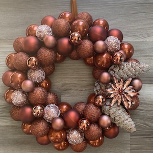 May include: A copper-colored Christmas wreath made of shiny, textured ornaments. The wreath is decorated with white, sparkly pine cones and a large, copper-colored star.