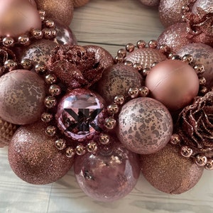 Peut inclure: Une couronne de No&euml;l rose gold compos&eacute;e d'ornements, de perles et de fleurs en tissu. Les ornements sont de diff&eacute;rentes tailles et textures, y compris des paillettes, du mat et du textur&eacute;.