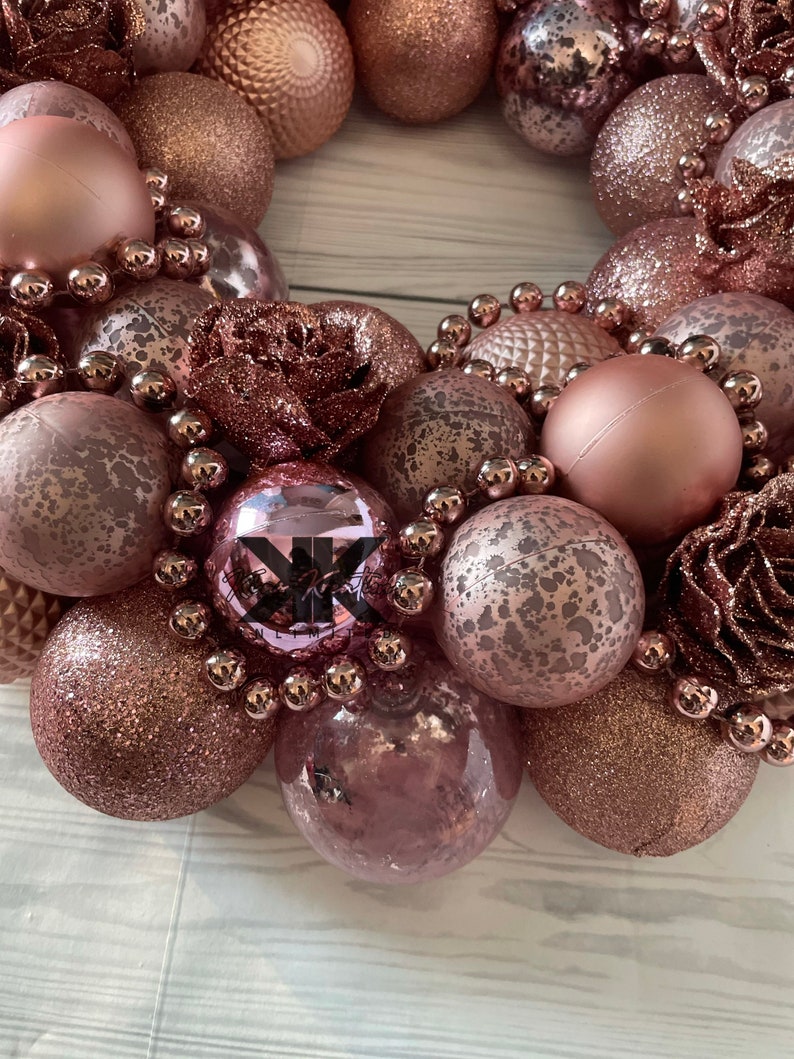 Peut inclure: Une couronne de No&euml;l rose gold compos&eacute;e d'ornements, de perles et de fleurs en tissu. Les ornements sont de diff&eacute;rentes tailles et textures, y compris des paillettes, du mat et du textur&eacute;.