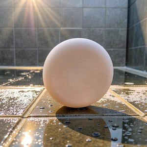 May include: A smooth, spherical, off-white bar of soap positioned upright. The surface of the soap is matte and uniform in color, contrasting with the surrounding dark, wet, tiled surface that reflects light and features scattered water droplets.