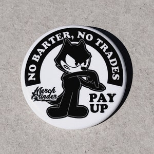 May include: Black and white button with a cartoon cat and the text "No Barter, No Trades Pay Up" and a logo that says "Merch Grinder".