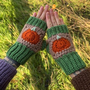 May include: A pair of handmade crochet fingerless gloves. The gloves feature a green cuff and a pumpkin design in orange and tan. The gloves are displayed against a background of green grass and sunlight.