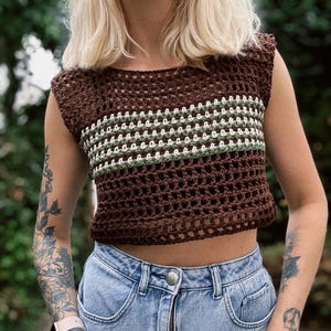 May include: A brown crochet crop top with a horizontal stripe design in white and green. The top is sleeveless and has a round neckline. The top is paired with light blue denim jeans.