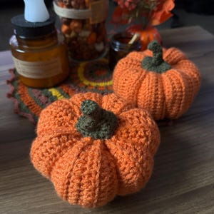 May include: Two crocheted orange pumpkins with green stems. The pumpkins are sitting on a wooden surface.