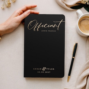 May include: A black notebook with gold lettering that reads "Officiant Annie Francis". The notebook also includes the names "Sarah & Tyler" and the date "08.08.2027". A gold pen and a cup of coffee are next to the notebook.