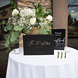 Black wedding guest book with gold foil text displayed on a reception table with elegant floral decor. Ideal for capturing guest messages, creating a personalized wedding keepsake, or adding a sophisticated touch to any wedding or special event.