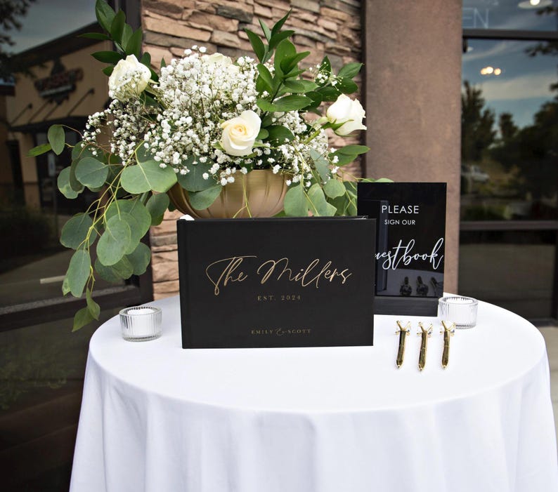 Black wedding guest book with gold foil text displayed on a reception table with elegant floral decor. Ideal for capturing guest messages, creating a personalized wedding keepsake, or adding a sophisticated touch to any wedding or special event.