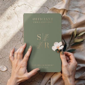 Close-up of a person's hands holding a personalized olive green officiant book with elegant gold foil lettering. The cover features "OFFICIANT Emma Johnston" and a stylish minimalist monogram with the couple's names and wedding date. Styled with a fresh white flower and olive branch on a rustic linen background. This custom ceremony notebook is a professional gift for a pastor or celebrant and a sophisticated keepsake for a modern, earthy-themed wedding. Classic officiant notebook