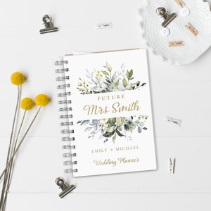 May include: A white wedding planner with a gold spiral binding. The cover features a watercolor illustration of green and gray leaves with the text "Future Mrs. Smith" in gold lettering. The text "Emily + Michael Wedding Planner" is also in gold lettering.