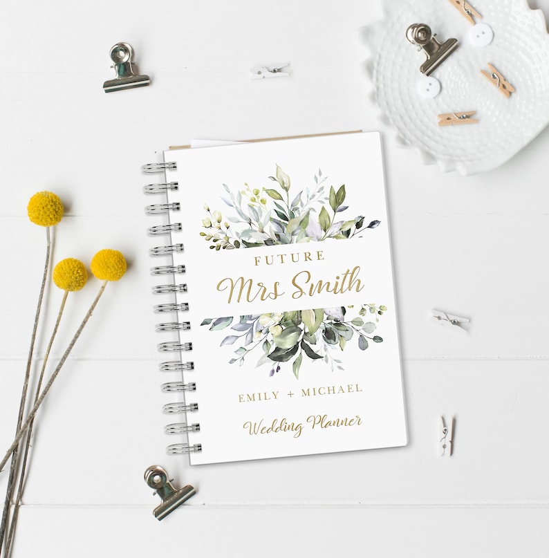 May include: A white wedding planner with a gold spiral binding. The cover features a watercolor illustration of green and gray leaves with the text "Future Mrs. Smith" in gold lettering. The text "Emily + Michael Wedding Planner" is also in gold lettering.