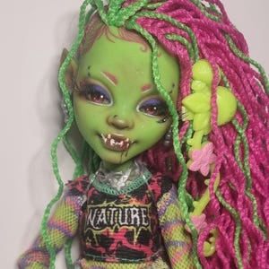 Peut inclure: Une poupée personnalisée avec une peau verte, des cheveux tressés roses et verts et un visage détaillé avec des crocs. La poupée porte un haut avec le mot "NATURE" et une ceinture argentée. Les bras de la poupée sont verts et la tenue comprend un jean bleu.