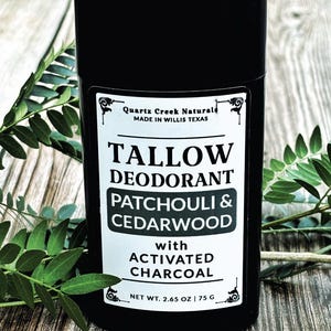 K&ouml;nnte beinhalten: Ein schwarzer zylindrischer Deodorant-Beh&auml;lter mit einem wei&szlig;en Etikett mit der Aufschrift Quartz Creek Naturals, Tallow Deodorant, Patchouli & Cedarwood with Activated Charcoal und Net Wt. 2.65 oz | 75 g. Das Produkt steht aufrecht neben gr&uuml;nen Bl&auml;ttern.